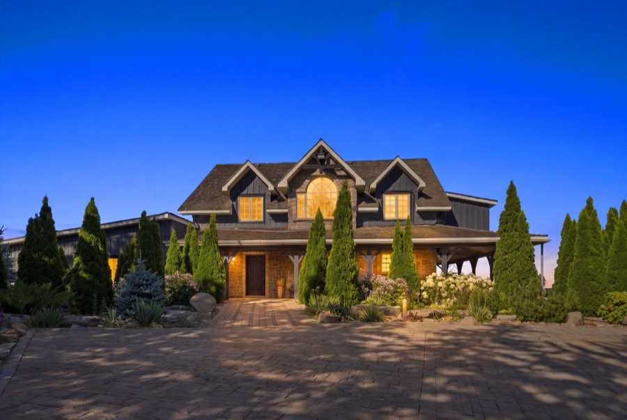 Front elevation of a gabled country home lit from within at blue hour, framed by tall cedars