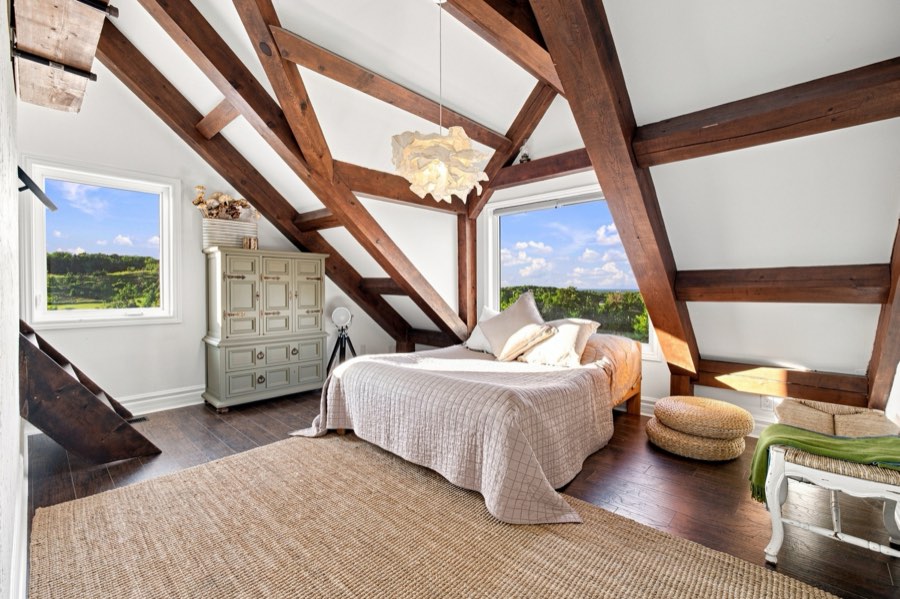 Loft bedroom framed by dark exposed trusses, with windows on two sides over open countryside
