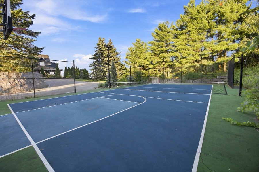 Fenced and floodlit blue sport court marked for tennis and basketball, ringed by evergreens